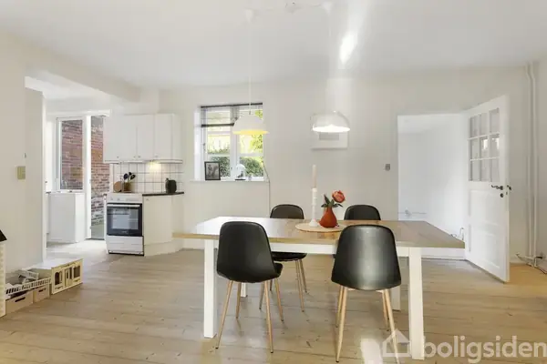 Spisebord med sorte stole står i lyst køkken-alrum med trægulv. Hvidt køkken med komfur og vindue ses i baggrunden. Rummets lysekrone hænger centralt over bordet.