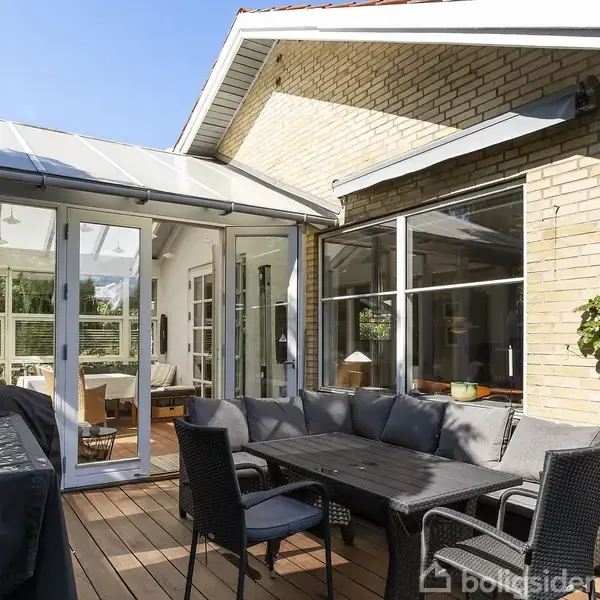 Terrasse med havemøbler inkluderer et sort bord og stole. Møblerne står på et trædæk ved siden af et glashus, der åbner mod en stue. Solen skinner klart.
