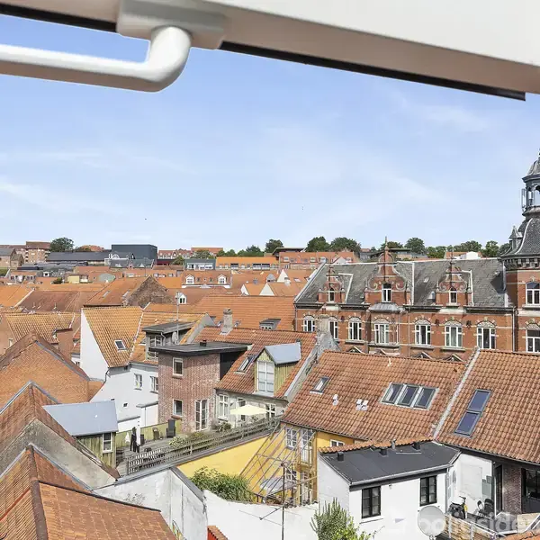 Udsigt over en by med huse med røde tage under en blå himmel. En kirkelignende bygning med klokketårn ses blandt hustagene, set fra et vindue med hvidt håndtag.