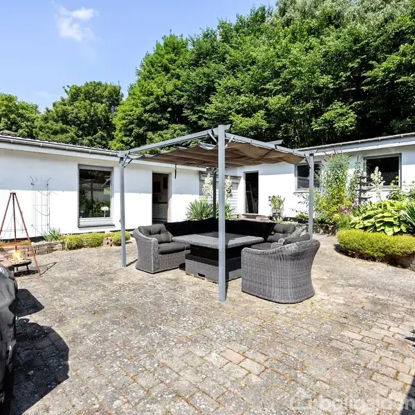 En gårdhave med fliser har en sort sofa- og stolgruppe under en pergola. Omgivet af en hvid bygning med vinduer, er der grøn vegetation i baggrunden.