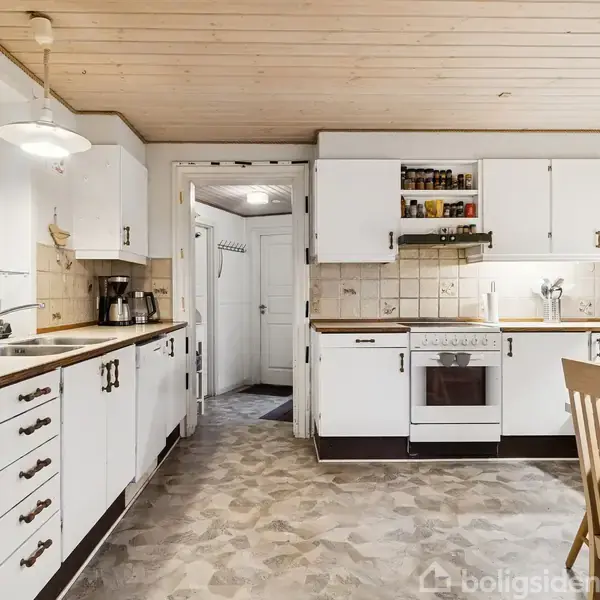 Køkken med hvide skabe og træbordplader, hvor en ovn og en kogeplade er indbygget. Til venstre er en vask, mens et spisebord står til højre. Vindue mod haven.