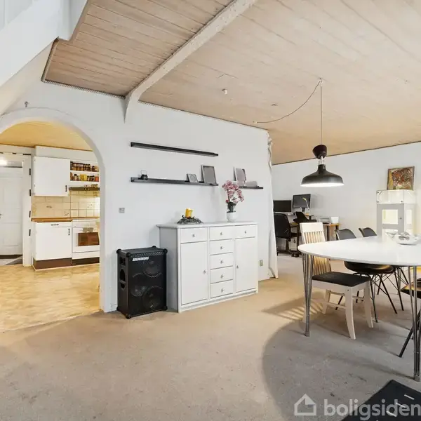 Spisebord med sorte stole står i en lys spisestue. En lampe hænger over bordet, omgivet af hylder og skænke. Bagved ses et hvidt køkken gennem en buegang.