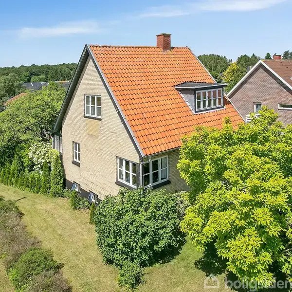 Et charmerende murstenshus med rødt tegltag står stille omgivet af frodige grønne buske. Det er placeret i et grønt boligområde med flere ensartede huse i baggrunden.