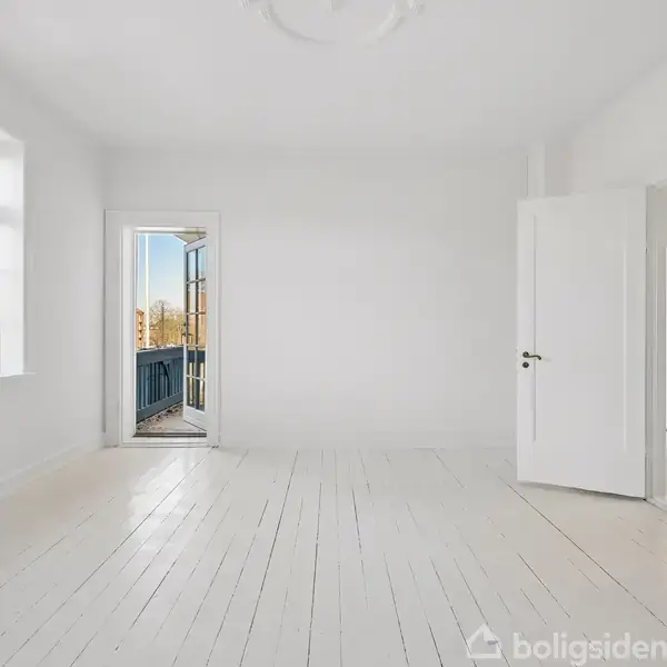 Et lyst og tomt værelse har hvide trægulve, en åben dør mod en blå balkon, og et stort vindue, der lader naturligt lys strømme ind.