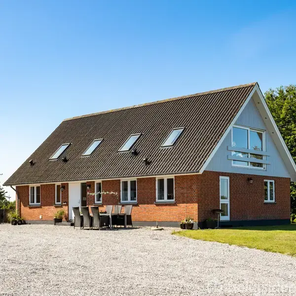 En rød murstensbygning med mørkt tag står stille omkranset af grønt græs og træer. Der er udendørsmøbler på grusarealet foran huset. Himmel er klar blå.