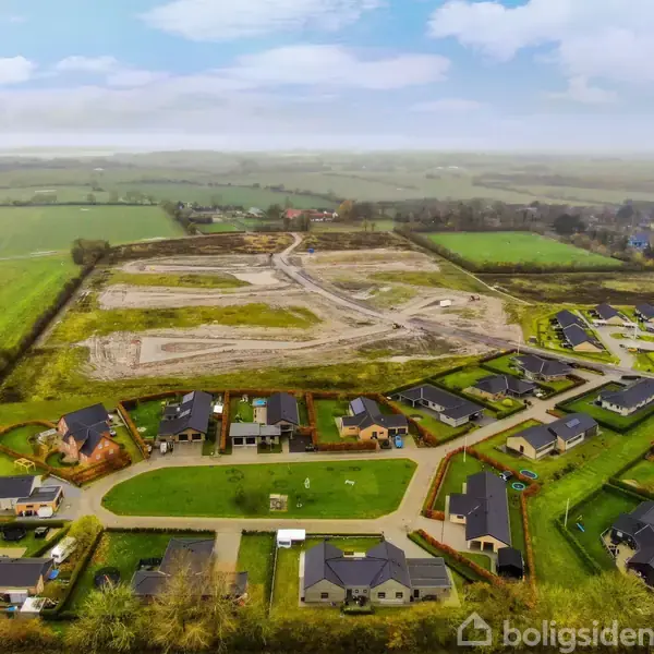 Luftfoto af en række moderne huse omgivet af grønne marker. En byggeplads med adskilte parceller ses i baggrunden. Området er roligt og naturskønt med spredte beboelsesområder.