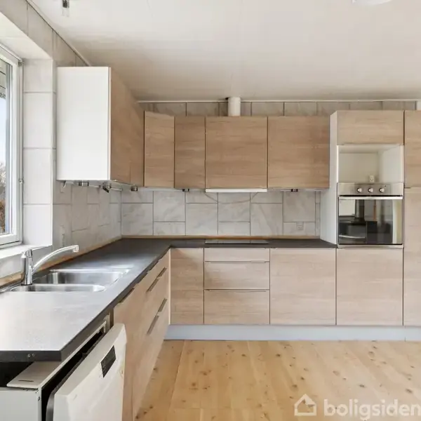 Køkken med moderne træskabe og indbygget ovn. Stålvask og opvaskemaskine på sort bordplade nær stort vindue. Hvidt køleskab i hjørnet, lyst trægulv understreger rummets lyse indretning.