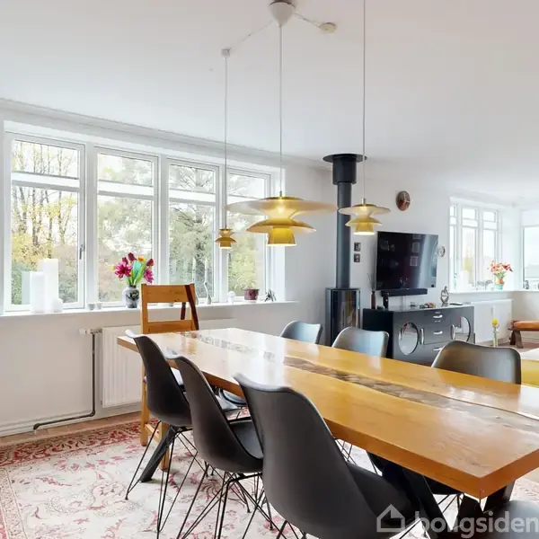 Langt spisebord med sorte stole står i en stue. Lamper hænger over bordet, og store vinduer slipper lys ind. En gul sofa ses ved siden af fjernsynet.