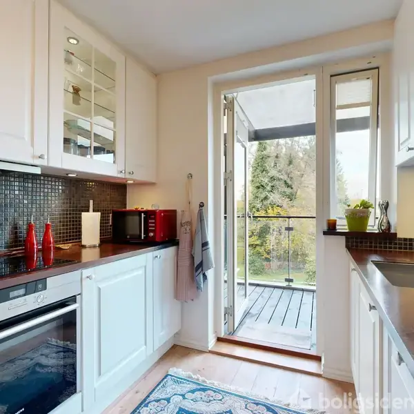 Et moderne køkken med hvide skabe og sort mosaikflise-bagvæg. En åbentstående glasdør leder ud til en altan med udsigt til træer. Rød brødrister og planter er på bordet.