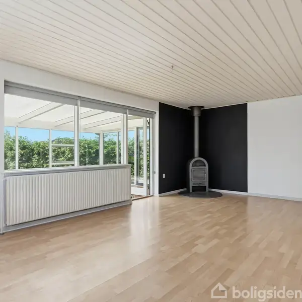 Stue med brændeovn mod en sort væg, store vinduer giver udsigt til en solrig terrasse og grønne træer. Lyse trægulve og hvidt loft fuldender rummets lyse udtryk.
