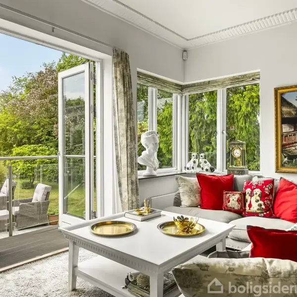 Stue med hvid sofa og røde puder står ved et vindue, der åbner ud til en terrasse med grøn udsigt. Hvidt sofabord med guldfade og billedramme på væggen.