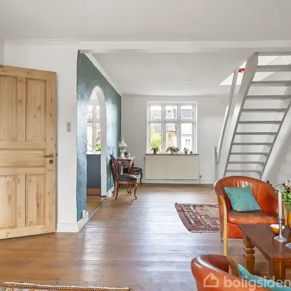 En stue med trægulv og hvidmalede vægge. En trappe fører opad, mens en trædør står åben. Rummet har siddeplads med puder og et bord med blomster. En bue fører til et vindue med udsigt.