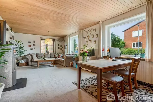 Et lyst stueområde med et stort hjørnesofaarrangement til venstre. Til højre står et bord med en bærbar computer. Vinduene giver udsigt til en have og nabohus.