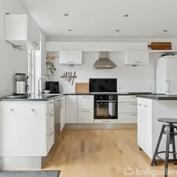 Et moderne køkken med hvide skabe og integrerede apparater. Køkkenet er rummeligt med en stor ø, omgivet af barstole. Vinduer bringer lys ind og planten udenfor er synlig.