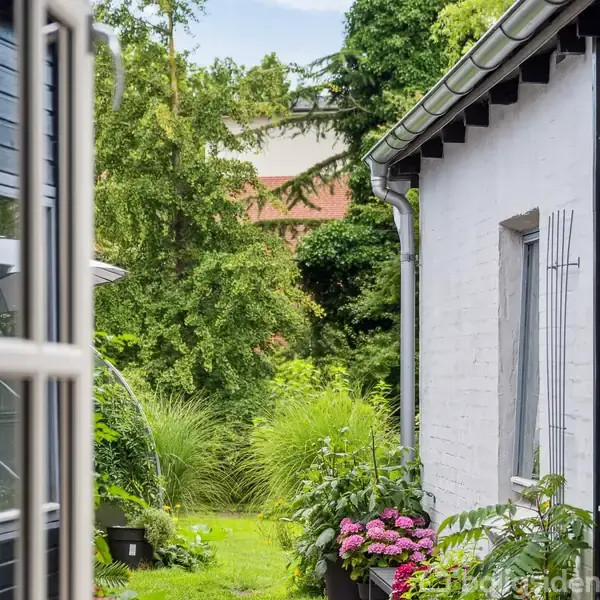 Røde murstenshuse står på en stille gade med parkerede biler og enkelte planter foran dørene. En blå himmel fylder baggrunden. Husene har karakteristiske skrå tage og hvide vinduer.