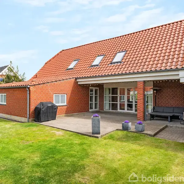 Et rødt murstenshus ligger stille i en grøn have, med en stor terrasse der rummer en grill og havemøbler. Taget er sat med flere Velux-vinduer, der slipper lys ind.