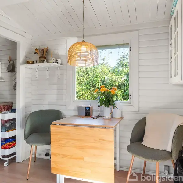 Et lille rum med træpaneler har et bord med to grønne stole og en blomsterbuket. Et vindue viser udsigt til grønne træer. Et køkkenområde med rødt gardin er i siden.