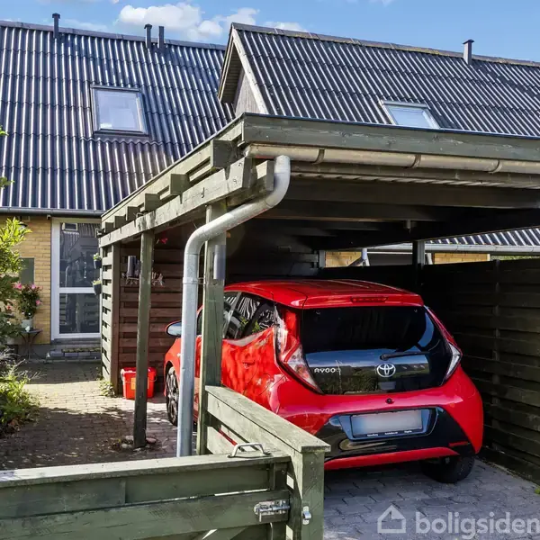 En rød Toyota Aygo holder parkeret under en carport ved et rækkehus med skråt, sort tag. Der er grønne planter ved siden af og flisebelagt indkørsel.
