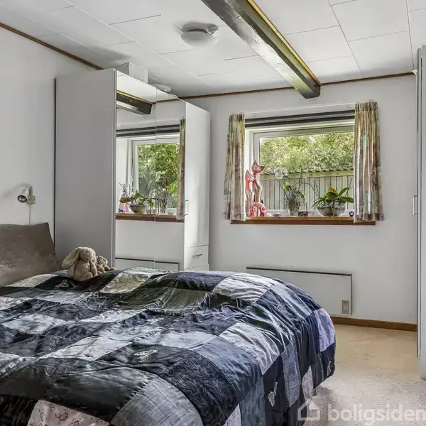 Soveværelse med en dobbeltseng og en farverig dyne, en bamse hvilende på sengen. Store vinduer med planter og pynt, skab med spejldør, kunstmaleri på væggen.
