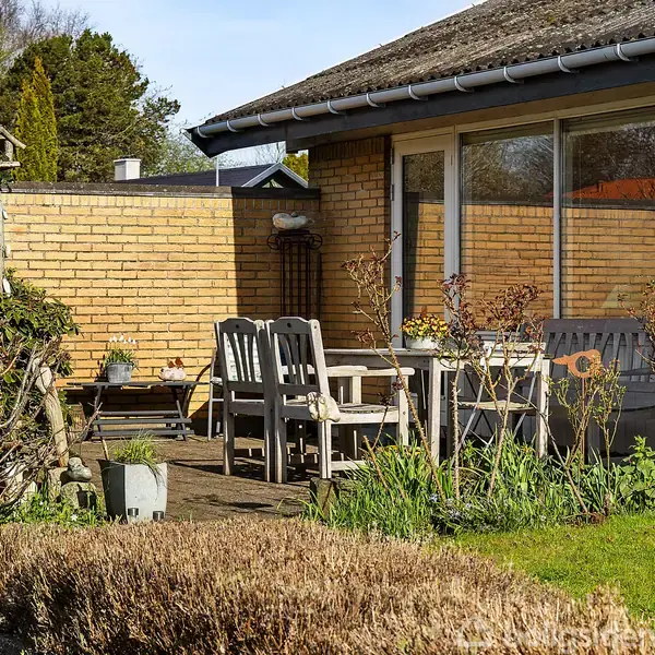 Et udendørs bord og stole sæt på en terrasse omgivet af frodig vegetation. Bygningen er af gule mursten med store vinduer, og omgivelserne ser rolige og velplejede ud.