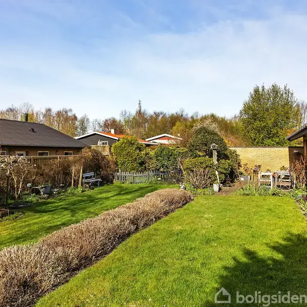 Grøn have strækker sig mellem to huse. En græsplæne er omkranset af buske og blomsterbed. I baggrunden ses flere træer, en terrasse og et bord med stole.
