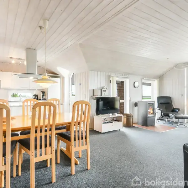 Et stort lyst opholdsrum viser et spisebord med højryggede stole, en sofa og en brændeovn. Et fladskærms-tv står ved siden af store vinduer, der fører ud til en terrasse.