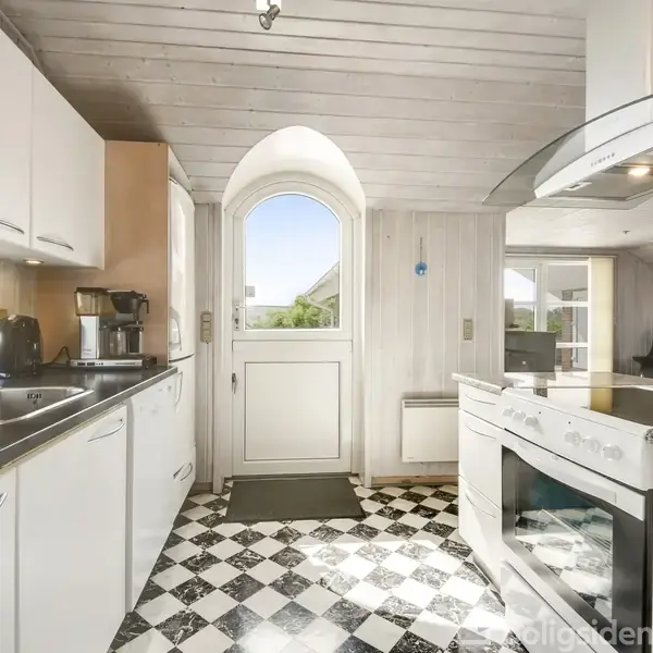 Køkken med hvide skabe og sorte bordplader, inkluderer komfur og vask. Store buede vinduer og en glasdør giver udsigt til en solrig have. Skakternet gulv i sort og hvid.