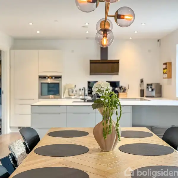 Et spisebord med sorte dækkeservietter står i et moderne, lyst køkken. En vase med blomster er centralt placeret på bordet. Køkkenet har hvide skabe og moderne apparater.