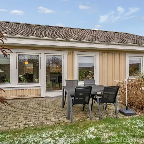 En enkelt etagers bygning med store vinduer, der viser et interiør med lamper, står omgivet af en flisebelagt terrasse med bord, stole og tæt bevoksning under en lyseblå himmel.