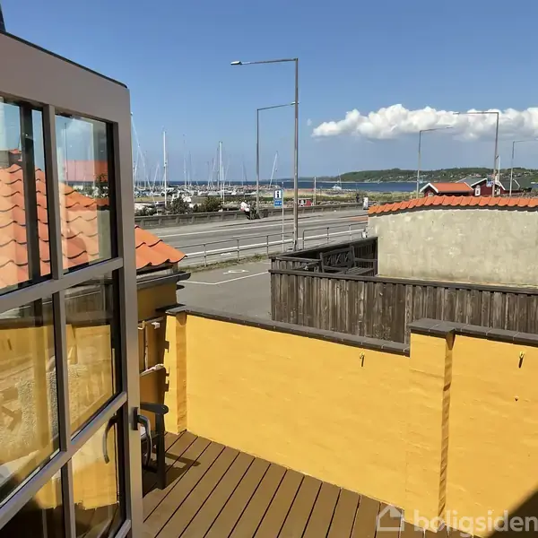 Udsigt fra balkon med gul mur og brune trægelændere, hvor man ser en vej og en havn med sejlbåde i baggrunden. Solrigt vejr og blå himmel over havn og vand.