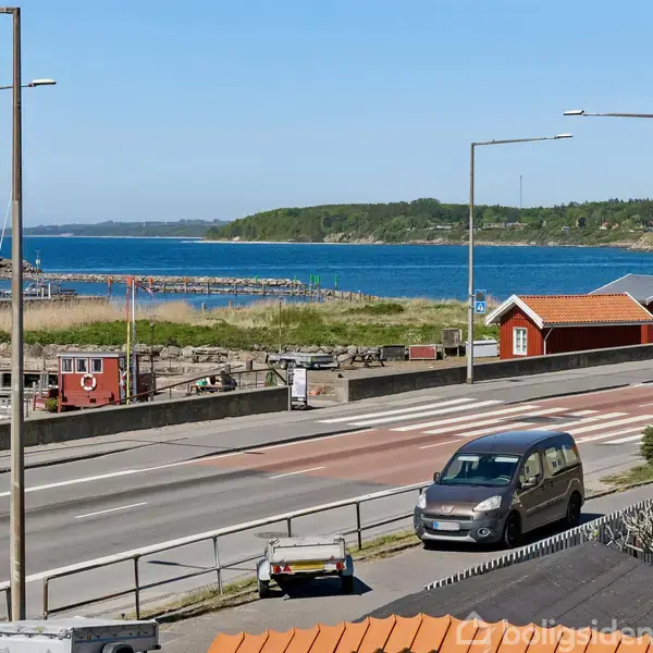 Biler kører på en vej langs en havn med små både. Havet er i baggrunden, og der er røde huse langs vejen. To vejskilte markerer information.