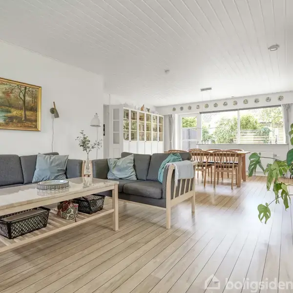 Sofaer med grå puder står i en lys stue med trælaminatgulv. Maleri hænger på væggen. I baggrunden ses et spisebord med stole og store vinduer med udsigt til have.