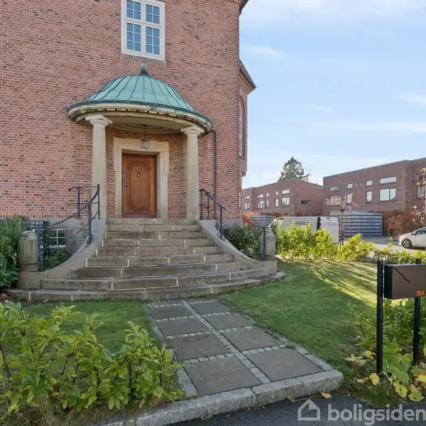 En murstensbygning med en rund indgangsportik med søjler og en grøn kuppelformet tag, hvor en trappe fører op til en trædør. Græsplæne og sti omgiver bygningen.