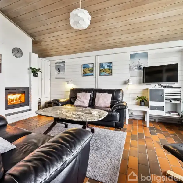 En stue med lædersofaer arrangeret omkring et rundt sofabord. En brændeovn opvarmer rummet. TV'et hænger på en hvid væg med kunstværker, og gulvet er flisebelagt.