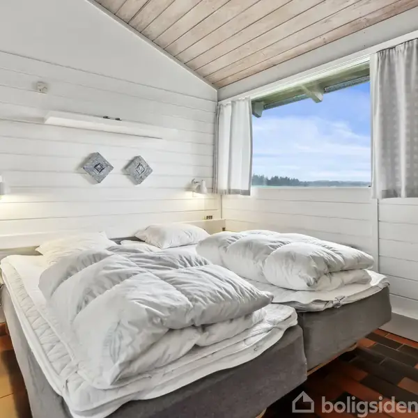En seng er dækket med hvide puder og dyner i et lyst soveværelse. Rummet har træpaneler, et vindue med gardiner og udsigt til natur udenfor.
