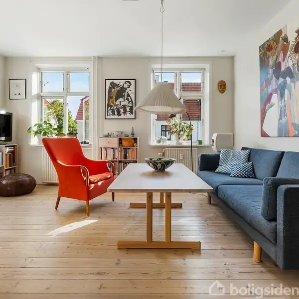 En stue med en blå sofa og rød lænestol omkring et træbord; væggene har kunstværker, og der er en bogreol og tv. Rummet er lyst med store vinduer.