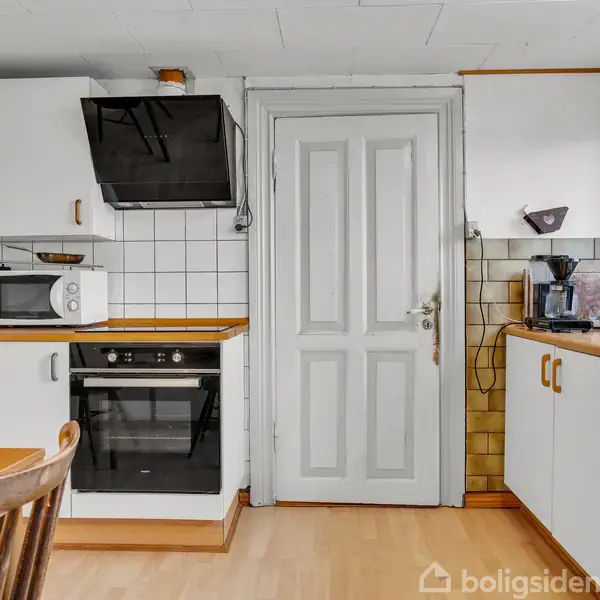 Et veludstyret køkken med ovn og mikrobølgeovn på en hvid bordplade, omgivet af hvide skabe. Kaffemaskine og elkedel står nær et vindue. Dør i midten, gul gardinkant.