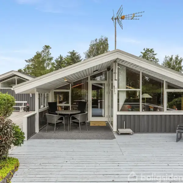Et sortmalet sommerhus med skråt tag har en stor, åben terrasse med havemøbler. Omgivet af træer og planter. Antenne på taget under en klar blå himmel.