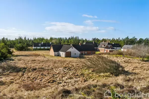 Flere huse står stille i et åbent, græsbevokset landskab omgivet af spredt skov. Himlen er blå og klar med få skyer.
