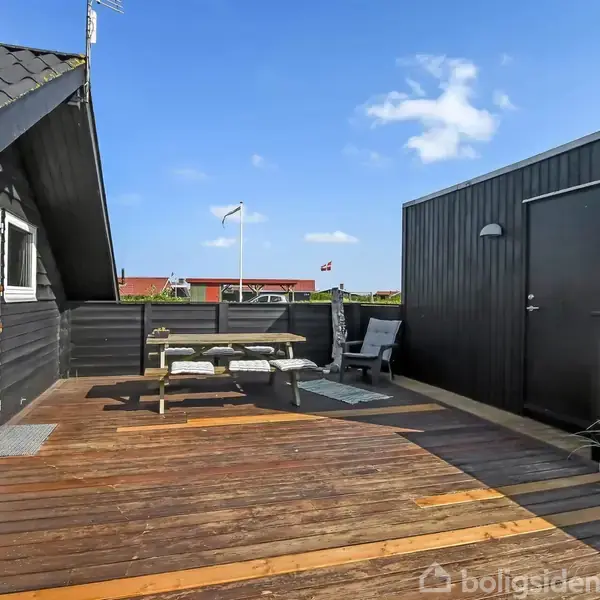 Et sort træhus med en stor træterrasse udstyret med et bord/bænkesæt og stole. En ting bygning i baggrunden under en skyfri blå himmel. En nummer 266 ses ved døren.