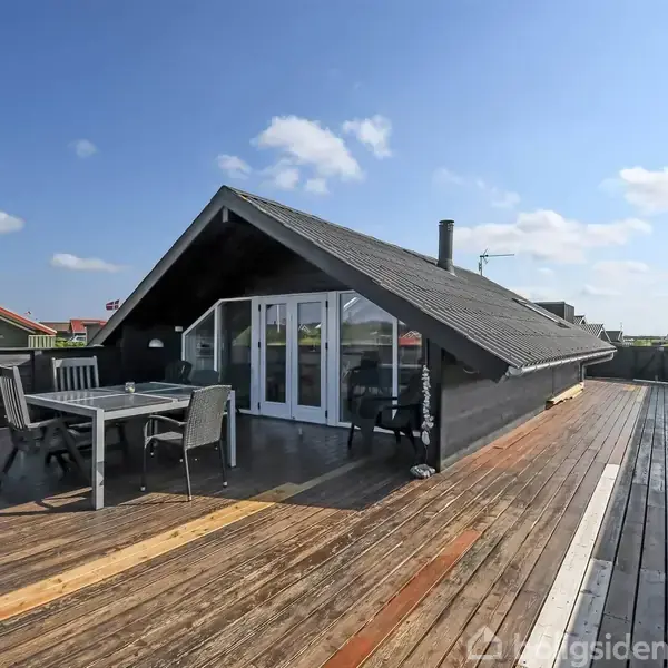 Mørkt sommerhus med skråtag har en træterrasse med møbler, herunder bord og stole. Blå himmel og naboer i baggrunden.