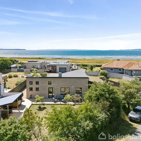 Et moderne hus med fladt tag står i en kystnær boligområde. Haven er velplejet, med udsigt til havet og en bil parkeret på vejen.