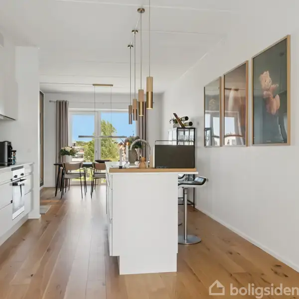 Et lyst køkkenalrum med træplankegulv, en køkkenø og moderne hængelamper. Langs væggen hænger der billeder. I baggrunden ses spisebordsområde med udsigt til træer udenfor.