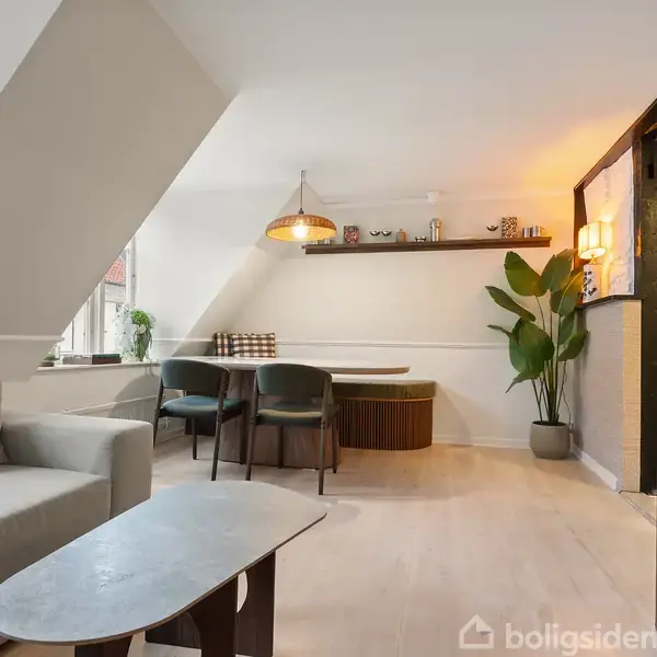 En moderne stue med en grå sofa og sofabord til venstre. Et spisebord med sorte stole står i hjørnet under en hængende lampe. En grøn plante og træmøbler fylder rummet harmonisk.