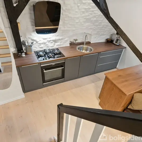 Køkken med mørkegrå underskabe og integreret ovn; træbordplade med gaskomfur og rund vask. Et træbord med stol på højre side. Omgivet af hvide vægge og synlige bjælker.