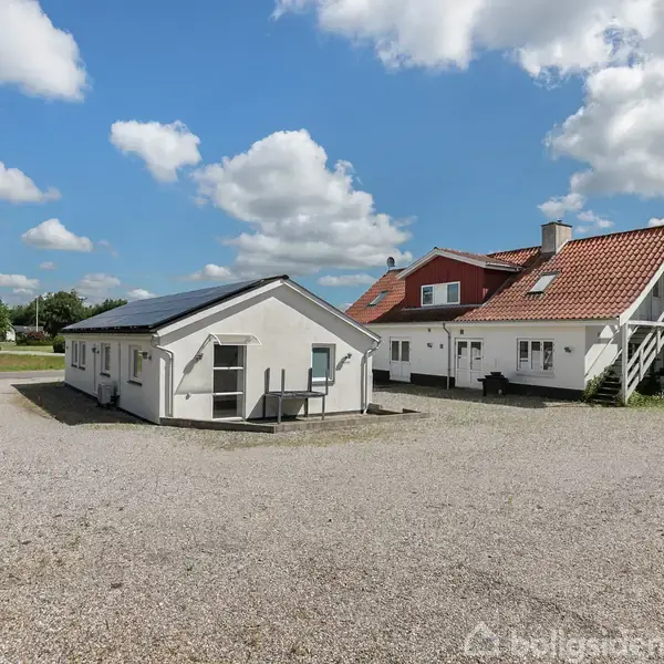 To huse står på en grusbelagt gårdsplads under en blå himmel med skyer. Det ene hus er hvidt med en rød tagstenstag. Det andet hus er lavere og hvidt.