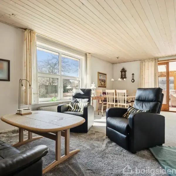 Stue med sorte lænestole og sofabord i midten. Store vinduer bader rummet i lys. Til højre står et spisebord med stole, omgivet af lyse gardiner.