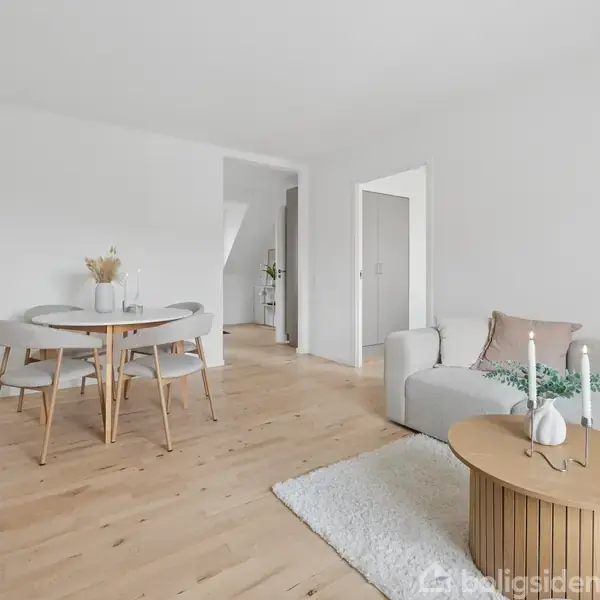 En lys stue med trægulve indeholder en grå sofa samt et rundt spisebord med stole. Store vinduer åbner ud til en balkon med udsigt til træer.