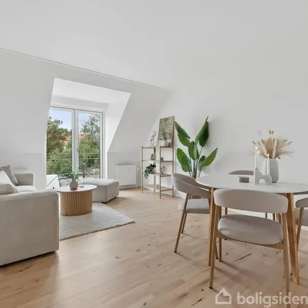 Et køkken med beige skabe og sort ovn integreret under kogeplade. Flere køkkenredskaber og planter står på bordet. Et vindue til højre giver udsigt over hustage.