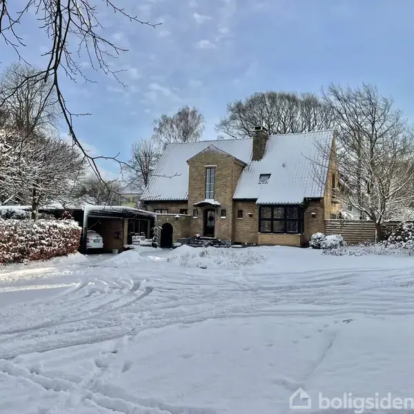Hus dækket af sne står omgivet af snedækkede træer og hække. En garage til venstre i billedet. Stemningen er vinterlig og rolig.